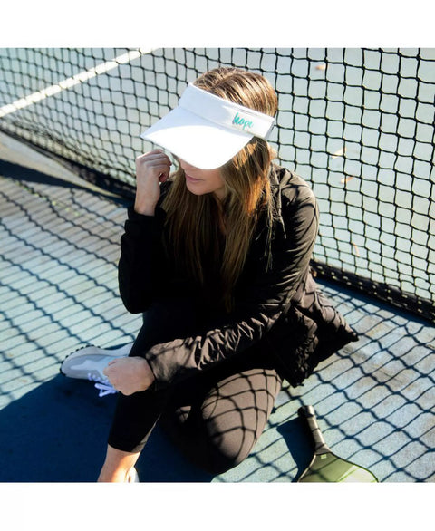 Person sitting on a tennis court wearing a visor and dark clothing.
