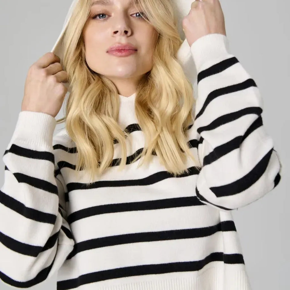 A blonde woman wearing a black and white horizontally striped knit sweater and a white wide-brim hat, posing against a solid gray studio background.