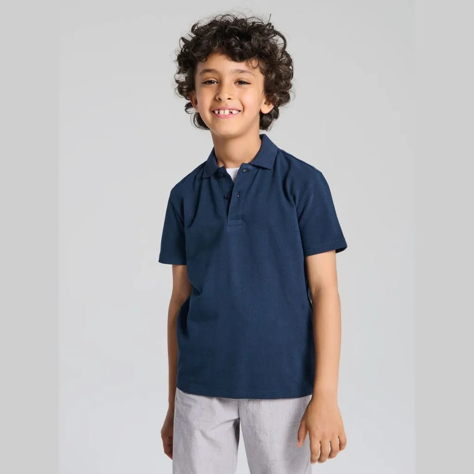 A young boy with curly dark hair smiling, wearing a navy blue short-sleeve polo shirt and light gray trousers, standing against a light gray studio background.