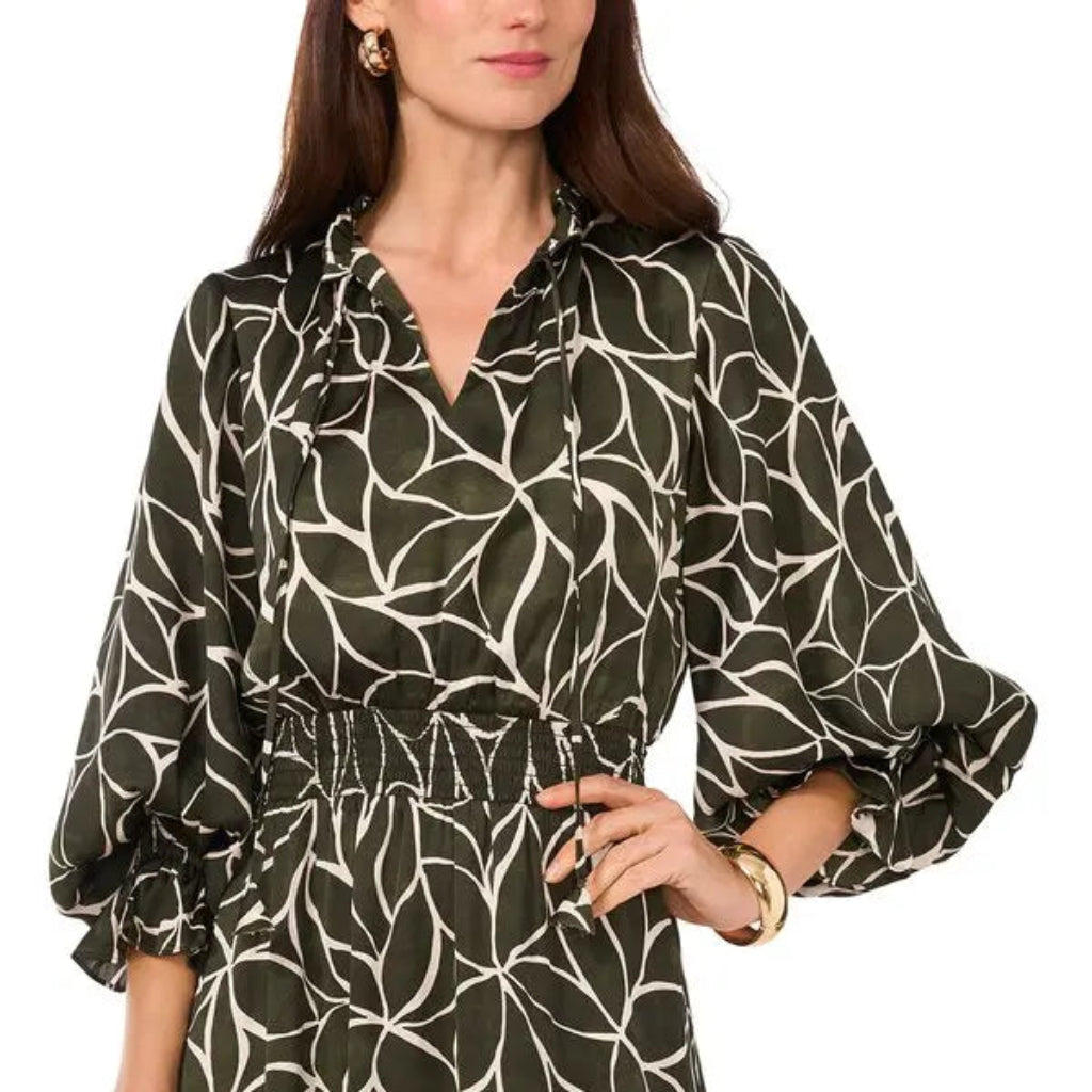 A woman wearing a dark olive green long-sleeve dress with a white abstract leaf-like pattern, featuring a tie-neck detail and a gathered elastic waist.