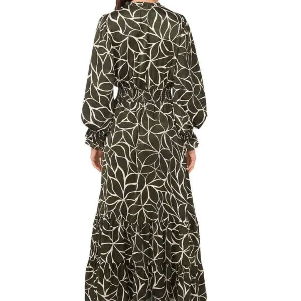 A woman viewed from the back wearing a long-sleeve maxi dress with a white abstract floral or leaf-like pattern on a dark olive green background, paired with black heeled boots against a plain white studio background.