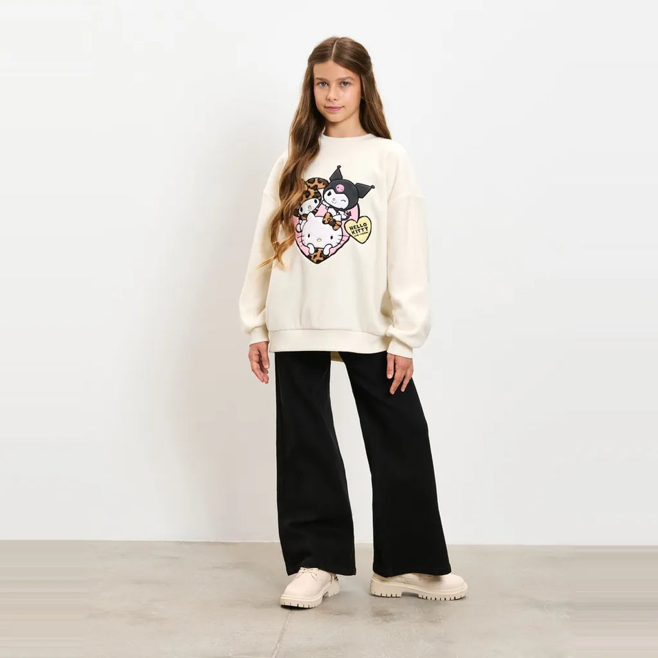 A young girl stands in a studio wearing a cream-colored sweatshirt with a colorful cartoon character graphic on the front, paired with black wide-leg pants and beige sneakers.