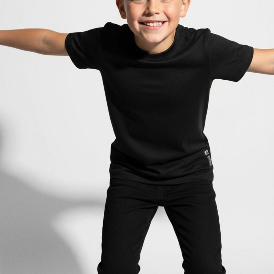 A smiling child wearing a black short-sleeve crewneck t-shirt and black pants, posing with arms outstretched against a light gray studio background.