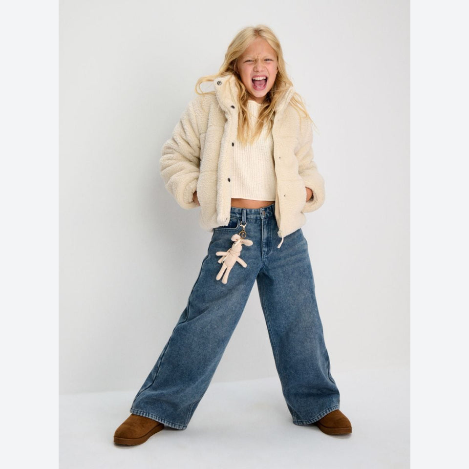 A young girl modeling a cream sherpa cropped jacket, cream knit top, wide-leg blue jeans with a plush keychain, and brown suede boots against a plain light gray studio background.
