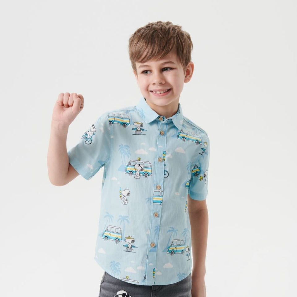 A young boy modeling a light blue short-sleeve button-up shirt with a cartoon animal print, paired with gray jeans featuring a Snoopy graphic, against a plain light gray background.
