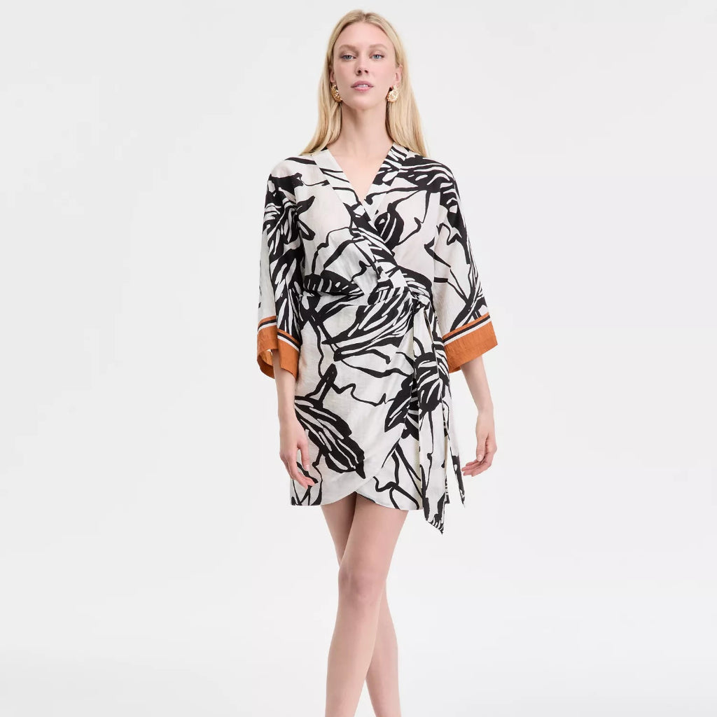 A woman modeling a short wrap-style dress with a black and white abstract floral print and contrasting orange cuffs, photographed against a clean white studio background.
