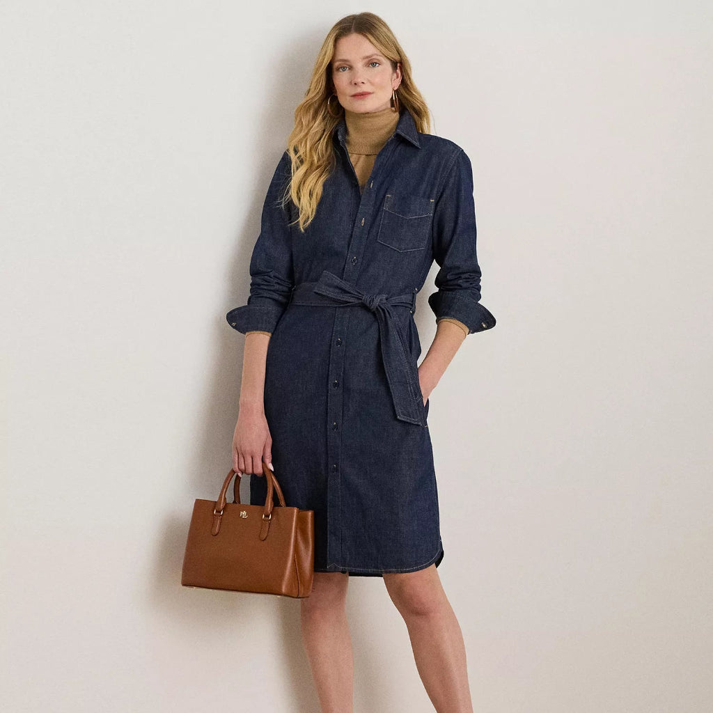 A woman models a dark blue denim shirt dress with a tie waist, paired with a tan turtleneck, brown leather handbag, and tan high heels, standing against a plain off-white wall.