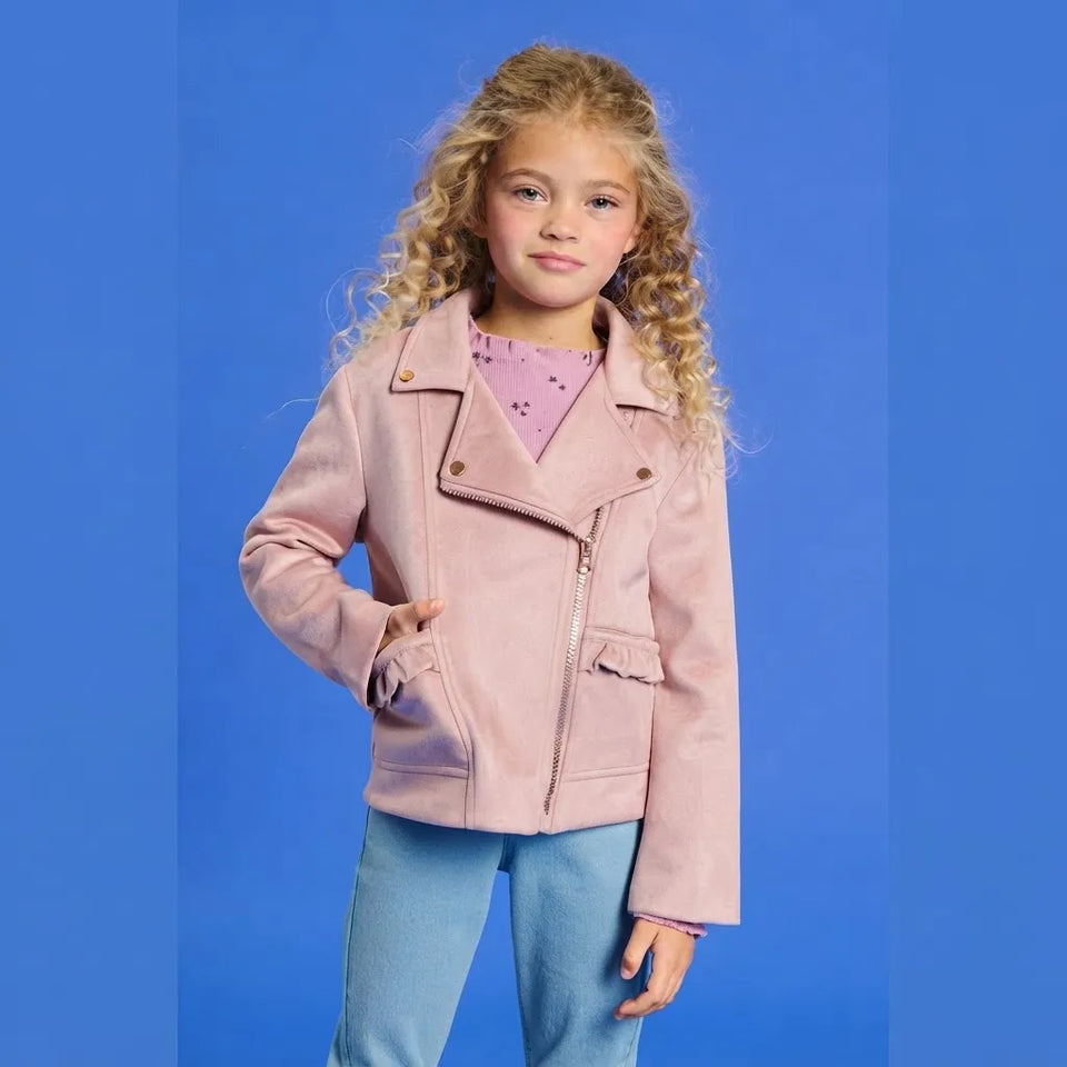 A young girl with curly blonde hair stands against a solid blue studio background wearing a light pink faux-leather moto jacket over a purple patterned top and light blue jeans.