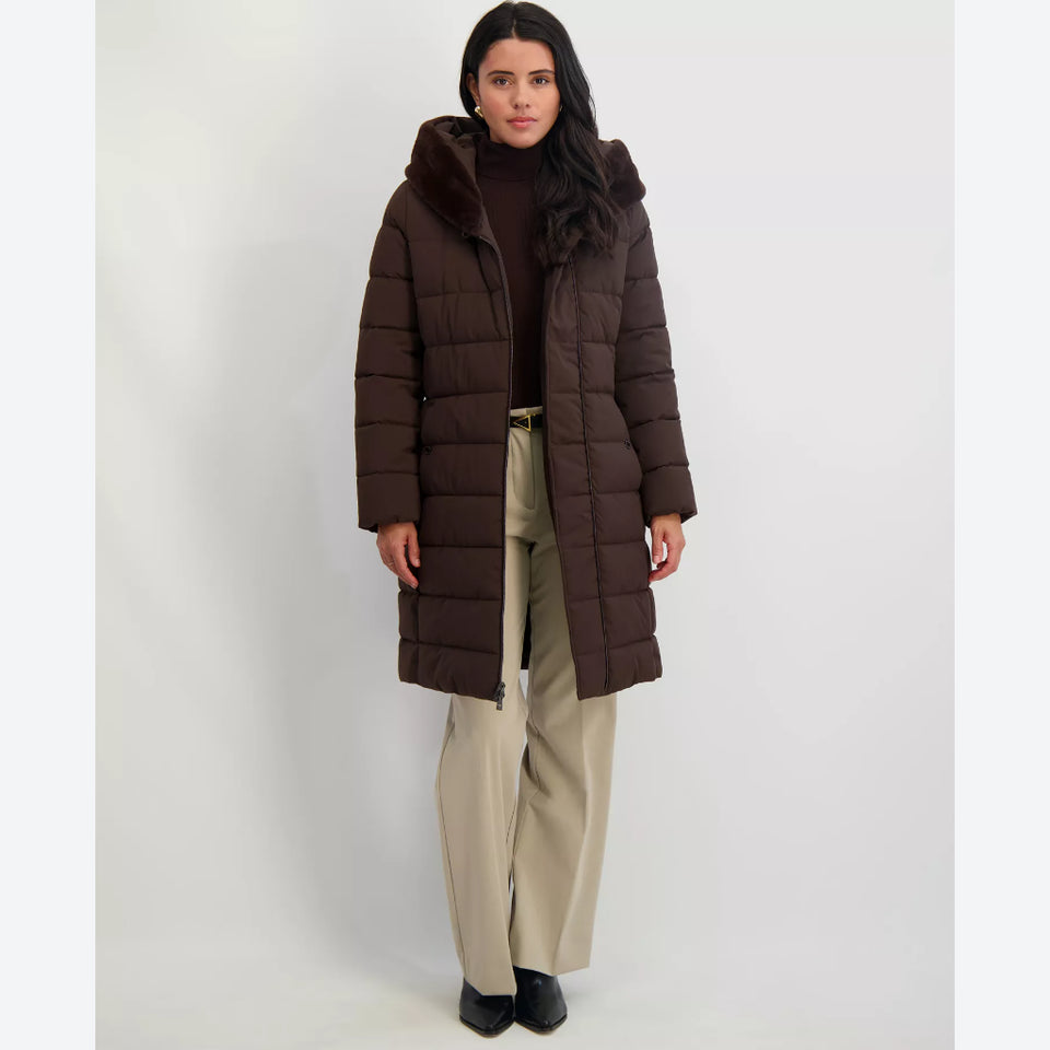 A woman modeling a long dark brown puffer coat with a large hood, worn over a dark top and paired with beige wide-leg trousers and black shoes, against a plain light gray studio background.