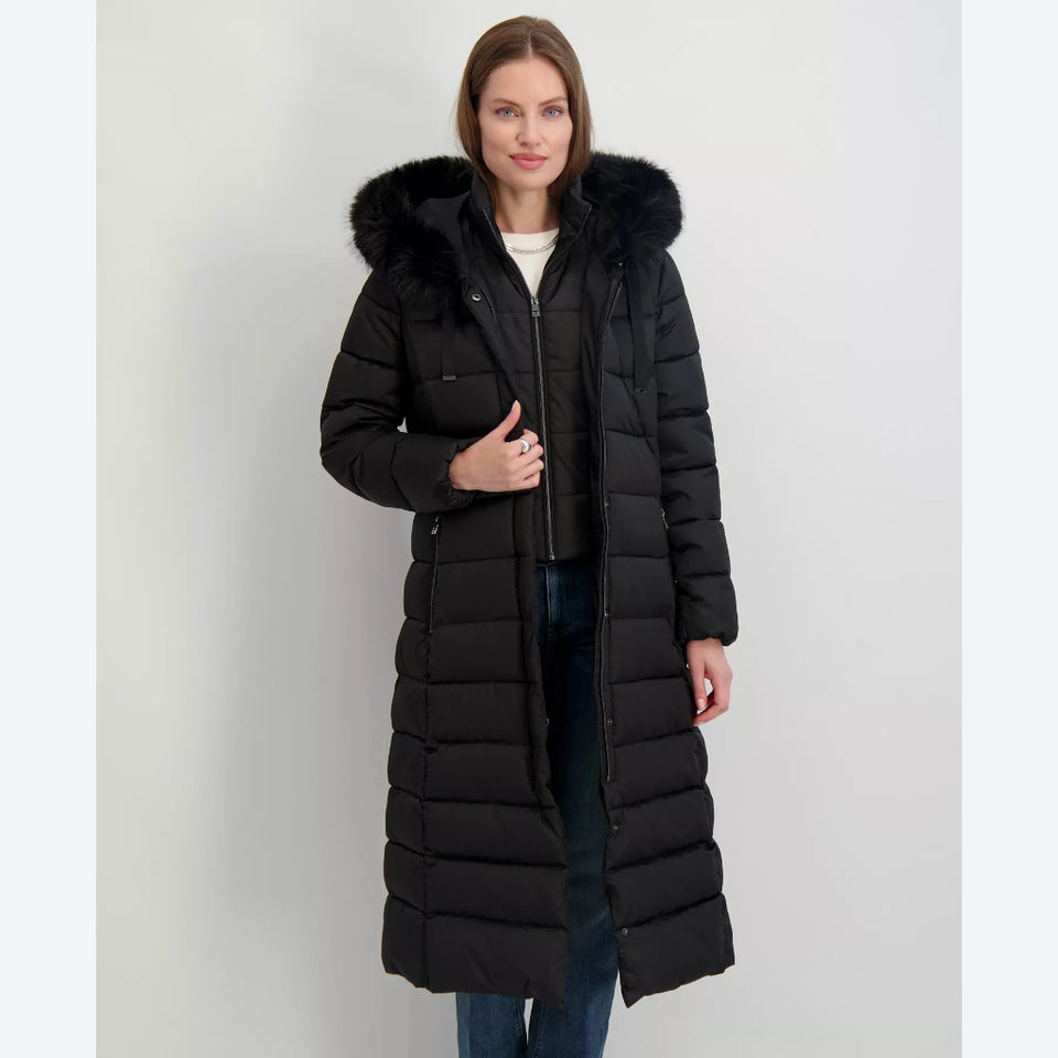 A woman modeling a long black puffer coat with a large black fur-trimmed hood, standing against a plain light gray studio background.