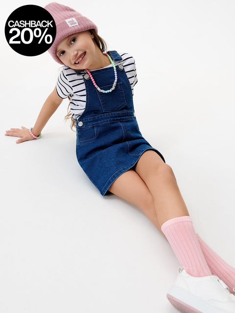 Child wearing a blue denim dress overalls, striped shirt, pink beanie, and socks on a white background