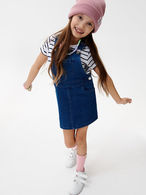 Young girl wearing a blue denim dress over a striped shirt with a pink beanie on a white background