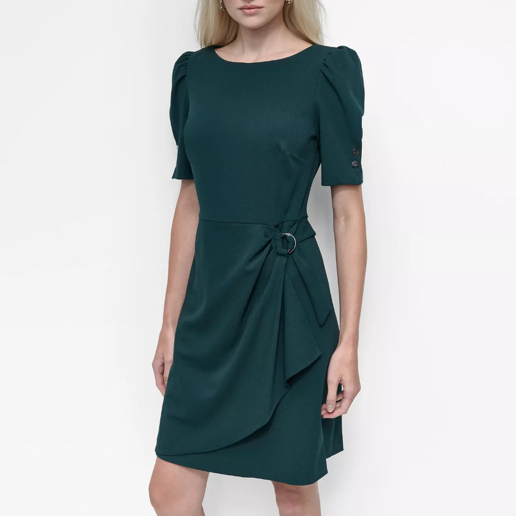 A blonde model wearing a dark green short-sleeve dress with puffed sleeves, a matching fabric belt with a round ring, and a draped wrap skirt, set against a plain white studio background.