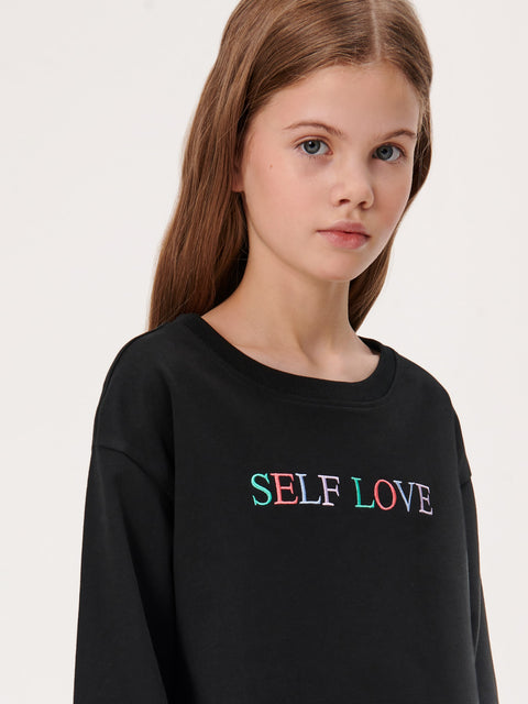 Young girl wearing a black sweatshirt with 'SELF LOVE' in colorful letters on a white background