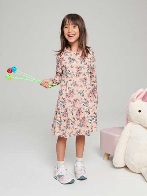 Young girl in a floral dress holding colorful sticks, standing next to a plush toy on a plain background