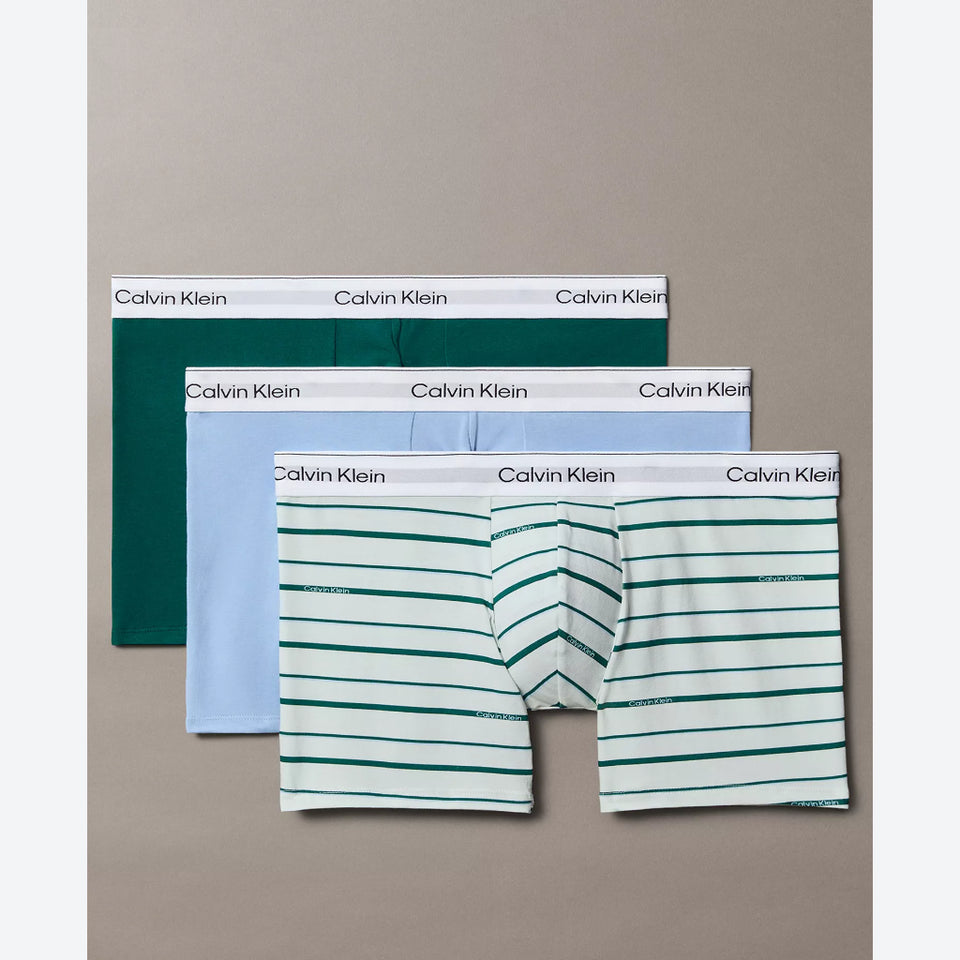 Three pairs of Calvin Klein men's boxer briefs arranged in a fan layout on a neutral taupe background. The front pair features a white base with thin green horizontal stripes, the middle pair is light blue, and the back pair is solid dark green.