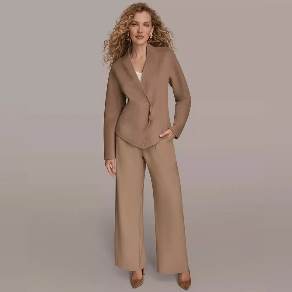 A woman modeling a matching taupe two-piece suit consisting of a structured wrap-style blazer and wide-leg trousers, paired with brown pointed-toe heels against a solid light gray studio background.