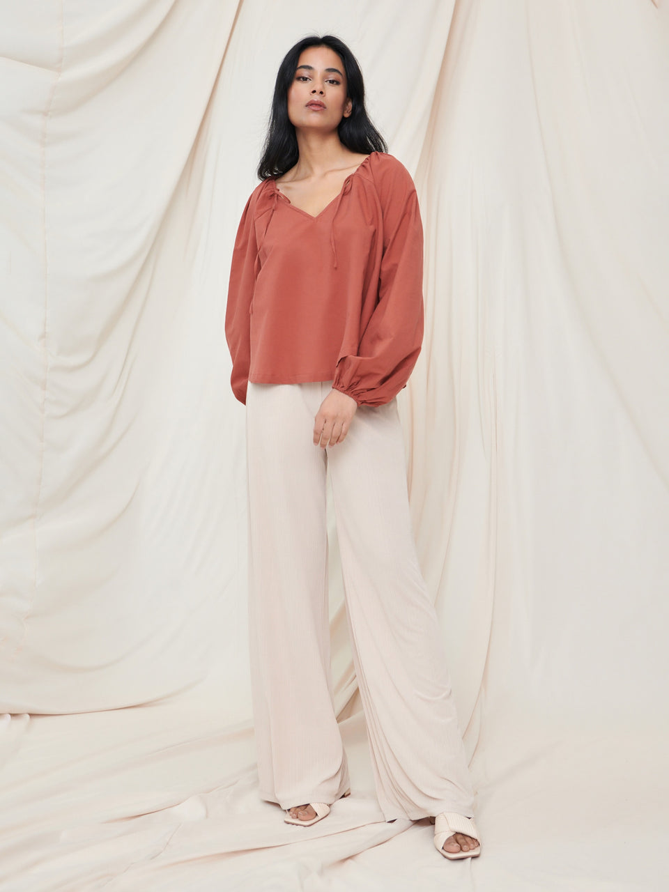 Woman wearing a rust-colored blouse and beige pants against a white curtain backdrop