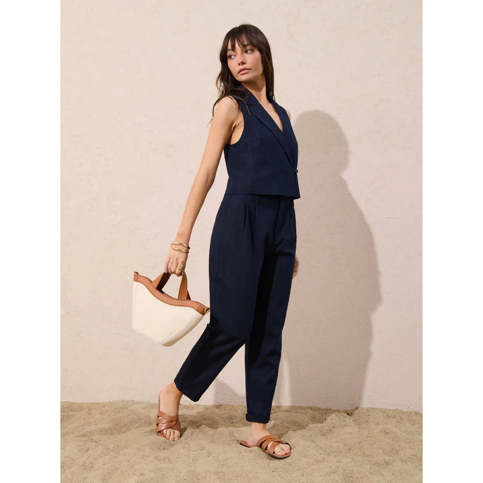 A woman models a navy blue sleeveless wrap-style jumpsuit with cropped pants, holding a cream and brown handbag against a textured beige wall and sandy floor.