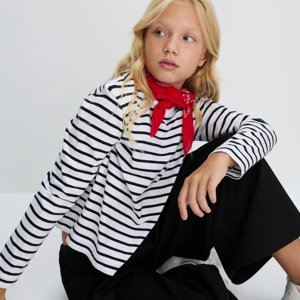 A young girl with blonde hair sits against a white background, wearing a black and white horizontally striped long-sleeve top, black wide-leg pants, and a red neck scarf.