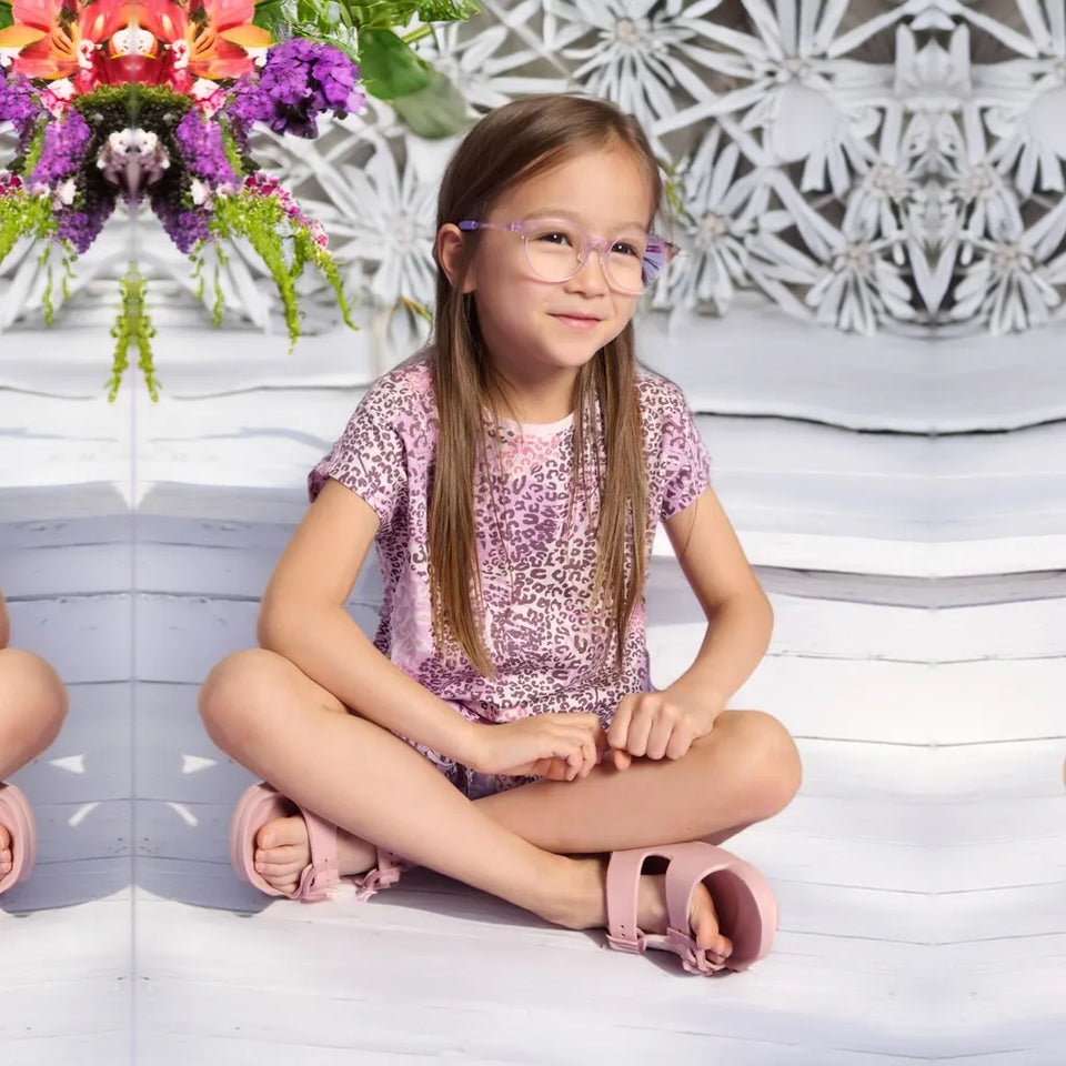 A young girl with long light brown hair sits cross-legged on white steps, wearing a pink leopard-print short-sleeve top, pink sandals, and pink-framed glasses. A mirrored floral arrangement is visible on the left side of the background.