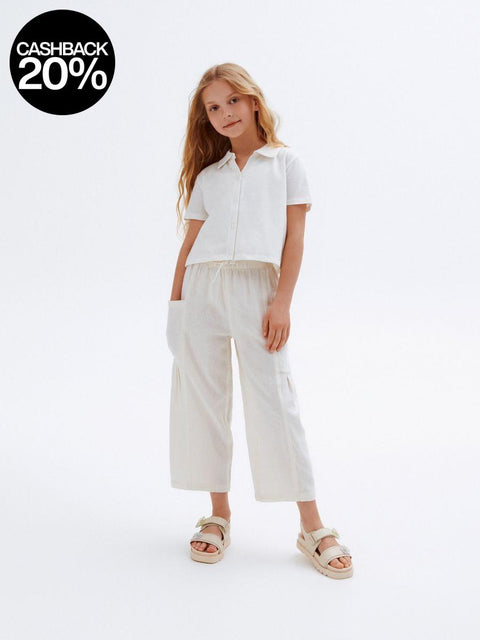 Young girl wearing a white shirt and beige pants on a white background with a cashback 20% badge.