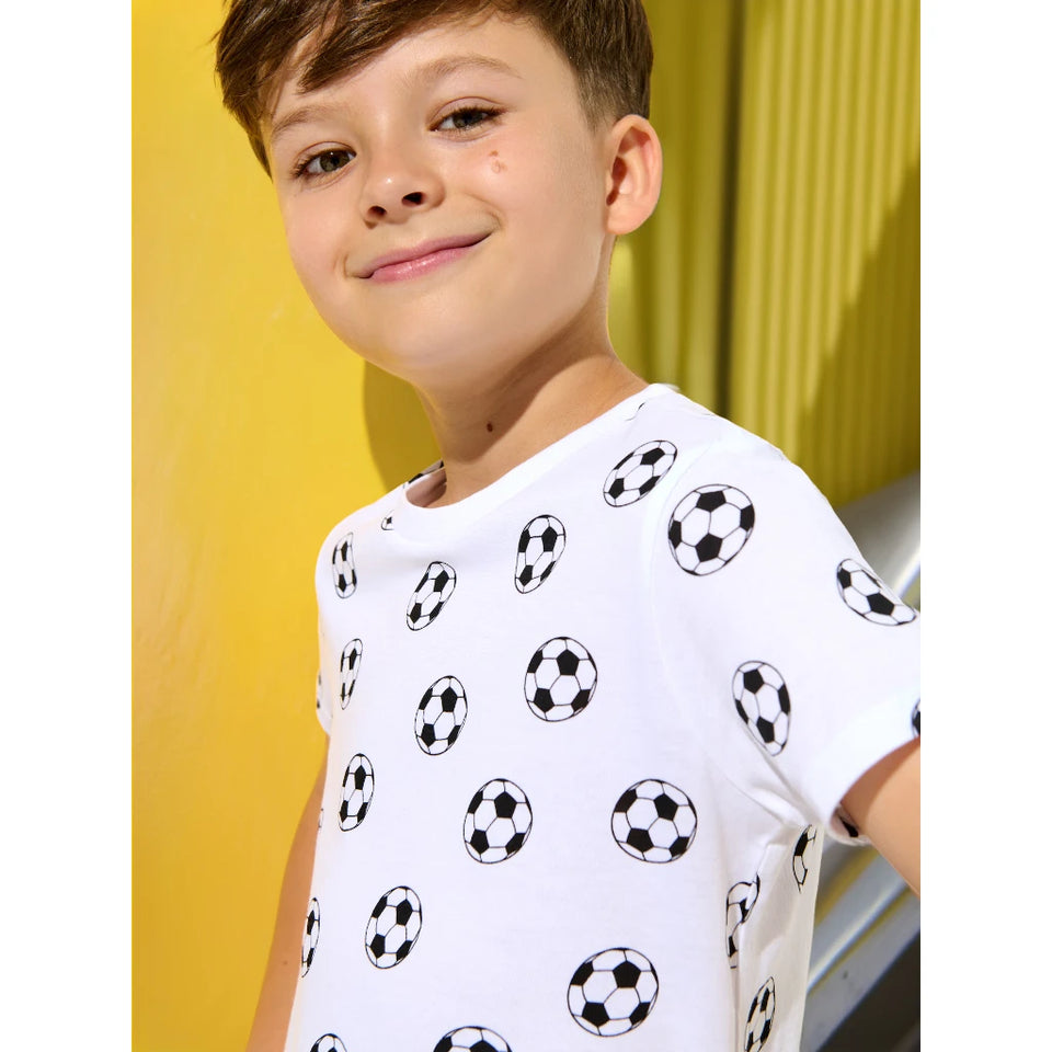 A young boy wearing a white short-sleeve t-shirt with a black and white soccer ball pattern, set against a bright yellow background.
