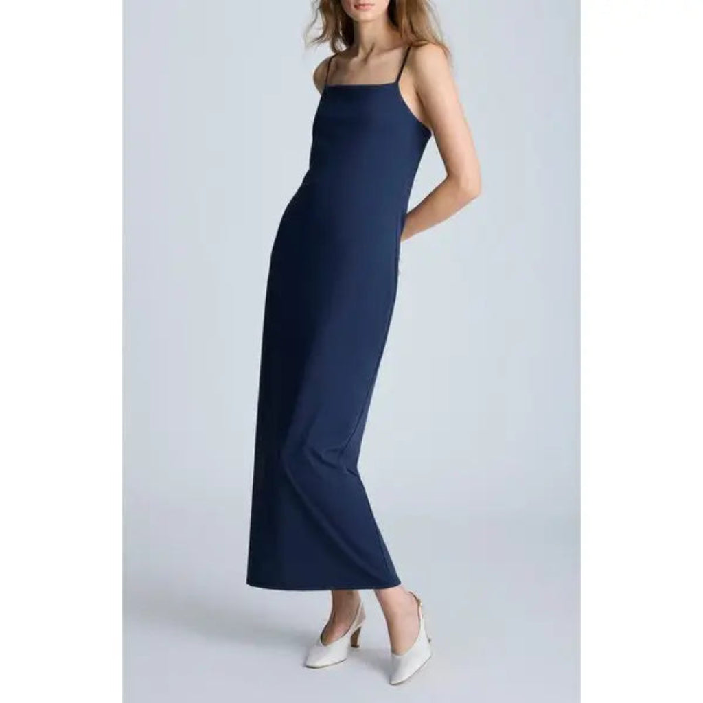 A woman modeling a navy blue maxi dress with thin spaghetti straps and a square neckline, paired with white pointed-toe heels, set against a light gray studio background.