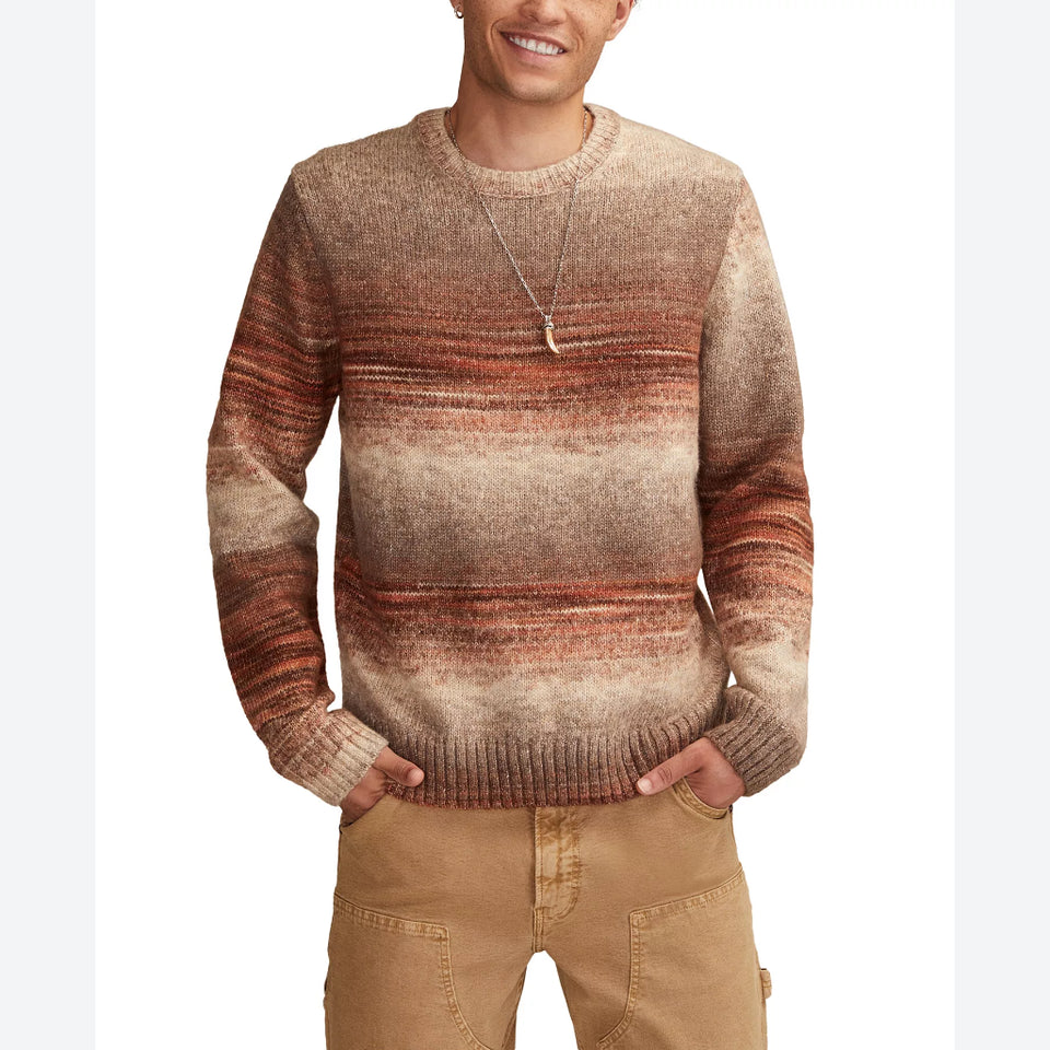 A man wearing a crewneck knit sweater with horizontal stripes in shades of brown, rust orange, and beige, paired with tan pants against a white background.