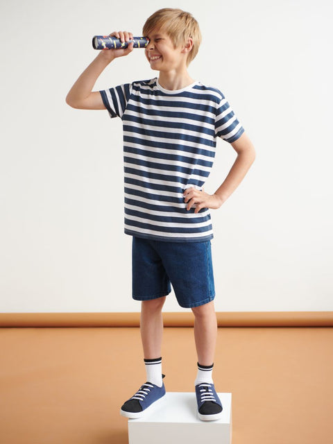 Young boy wearing a striped shirt and shorts, standing on a white box with a brown background.