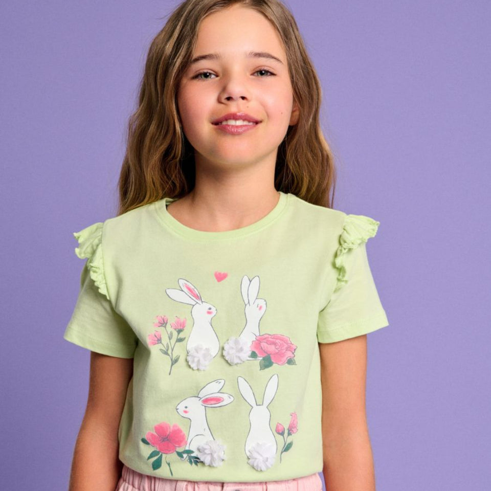 A young girl with light brown hair is wearing a light green short-sleeve t-shirt with a bunny and floral print, paired with pink pants, set against a solid purple background.