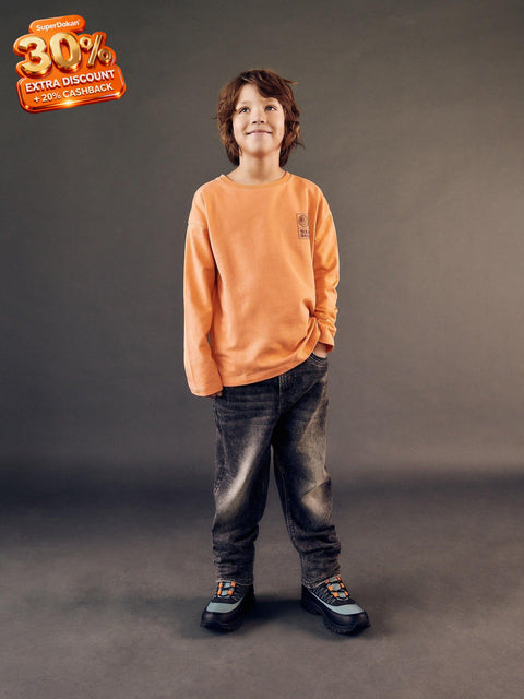Child wearing an orange sweatshirt and dark jeans standing against a gray background