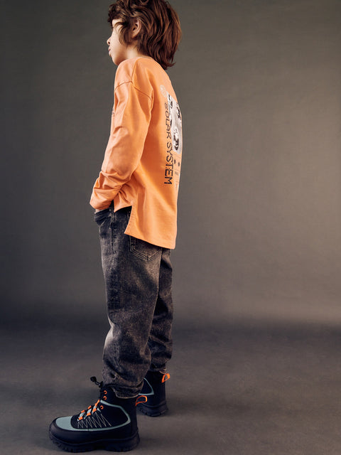 Child wearing an orange long-sleeve shirt and dark jeans standing against a gray background