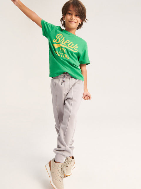 Child wearing a green t-shirt with text and gray pants on a white background