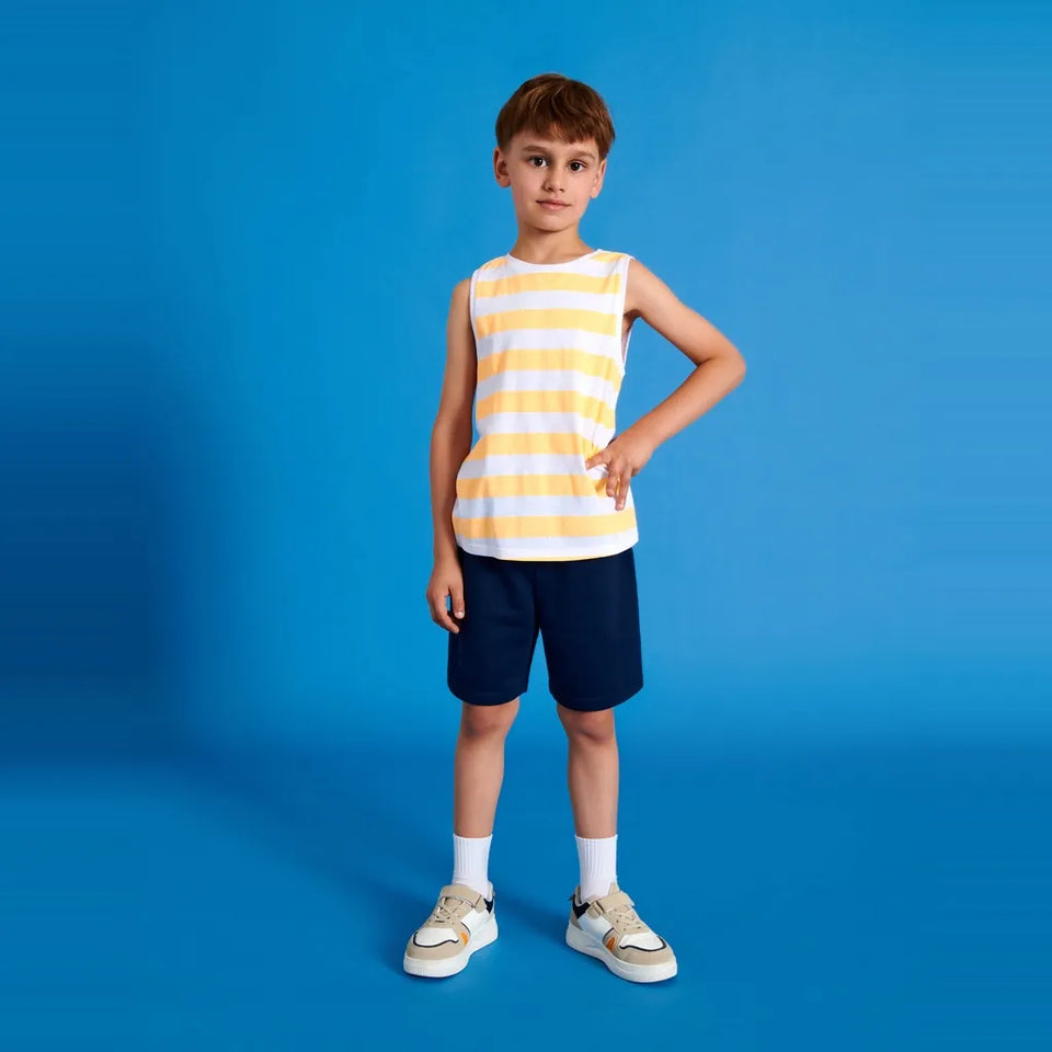 A young boy stands against a solid blue studio background wearing a sleeveless tank top with yellow and white horizontal stripes, navy blue shorts, white socks, and light-colored sneakers.