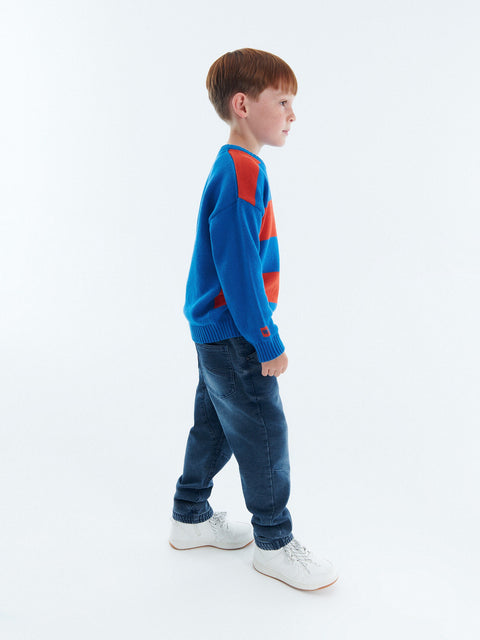 Young boy wearing a blue sweater with red accents and jeans, standing against a white background