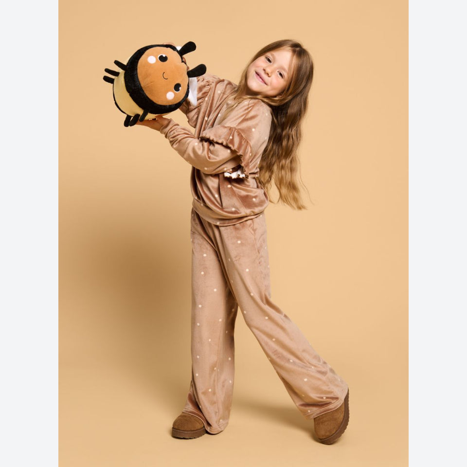 A young girl with long light brown hair stands in a studio wearing a matching beige pajama set with small white polka dots. She is holding a plush bumblebee toy against a solid warm beige background.