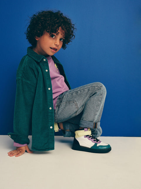 Child wearing a green jacket, purple shirt, and jeans with colorful sneakers against a blue background
