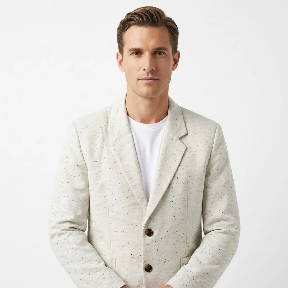 A man wearing a light beige textured blazer with small dark speckles over a white crewneck t-shirt, photographed against a clean white studio background.