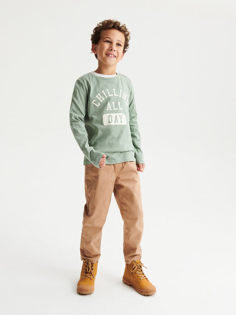 Child wearing a green sweatshirt with text, beige pants, and brown boots on a white background