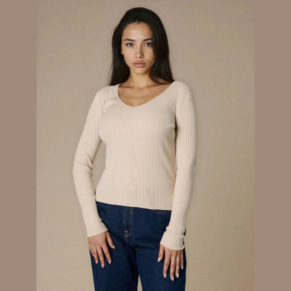A woman modeling a ribbed long-sleeve scoop-neck sweater in cream, paired with dark blue jeans, against a solid beige studio background.