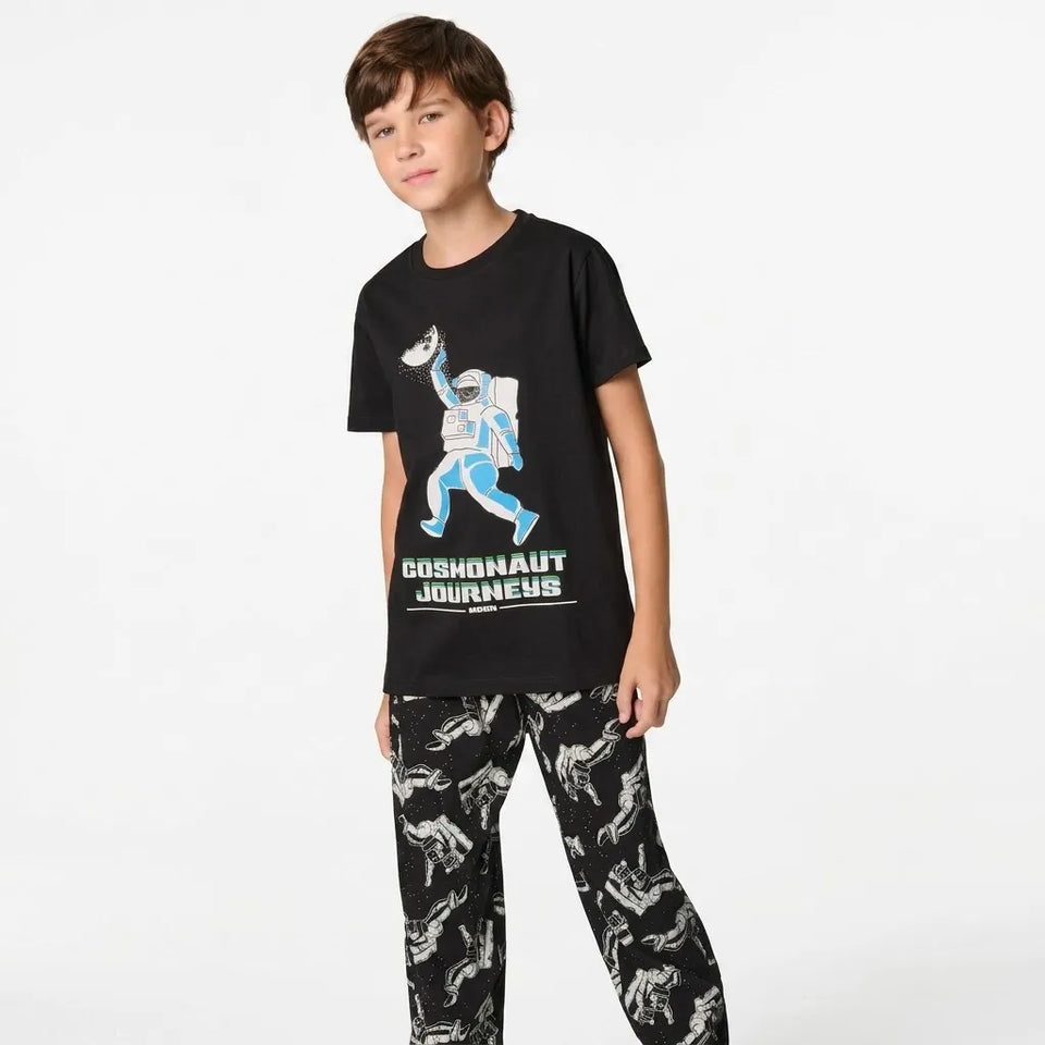 A young boy modeling a black short-sleeve t-shirt with a blue and white astronaut graphic and the text 'COSMONAUT JOURNEYS' paired with matching black pajama pants featuring a white space-themed dinosaur pattern, set against a plain white studio background.