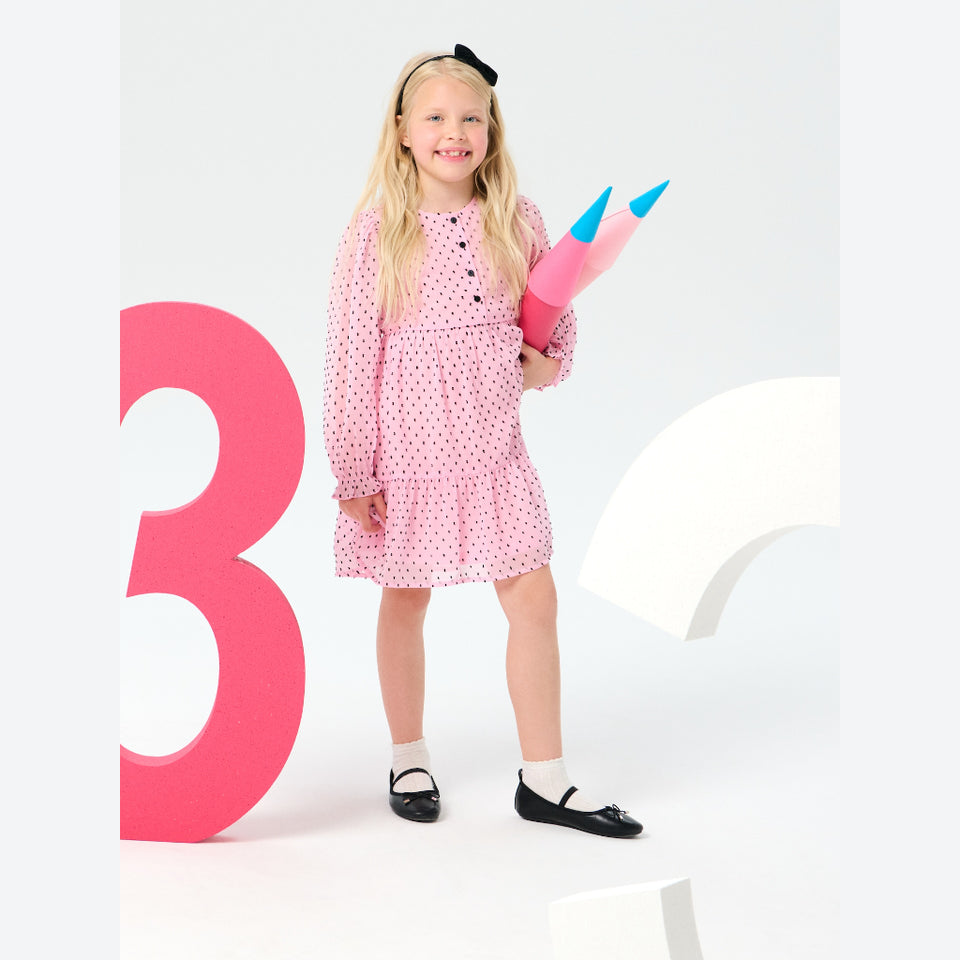 A young girl with blonde hair stands in a studio wearing a pink long-sleeve dress with small black polka dots, white socks, and black Mary Jane shoes. She is holding a large pink pencil-shaped plush toy.