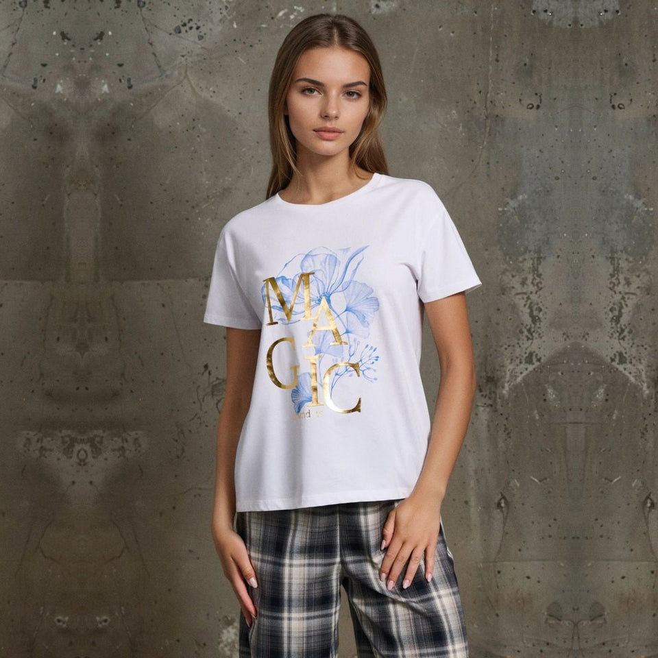 A woman modeling a white short-sleeve graphic T-shirt with a blue floral design and gold metallic 'MAGIC' lettering, paired with plaid pants, set against a textured gray concrete wall.
