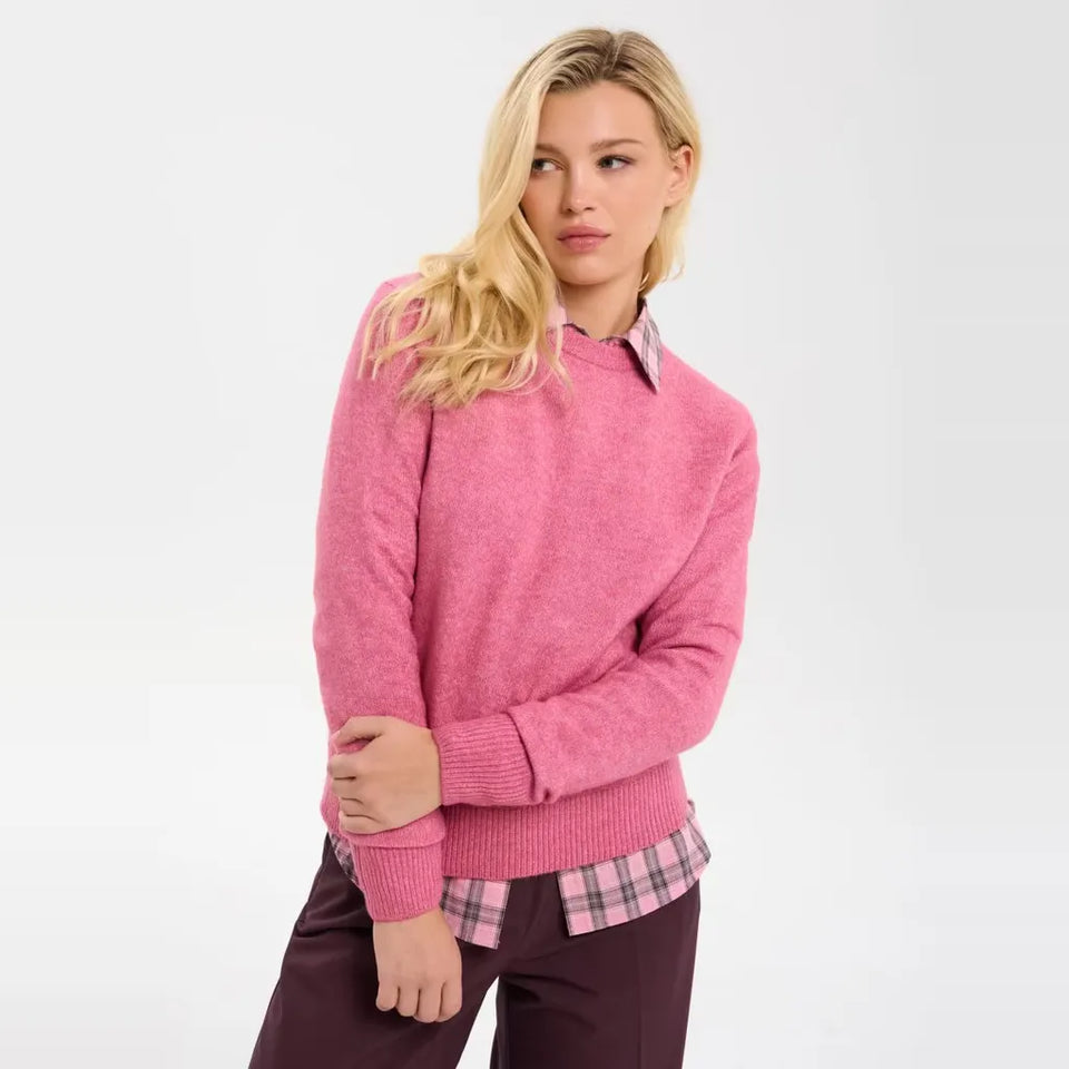 A blonde model wearing a pink knit mock-neck sweater over a pink plaid collared shirt and dark trousers, photographed against a clean white studio background.