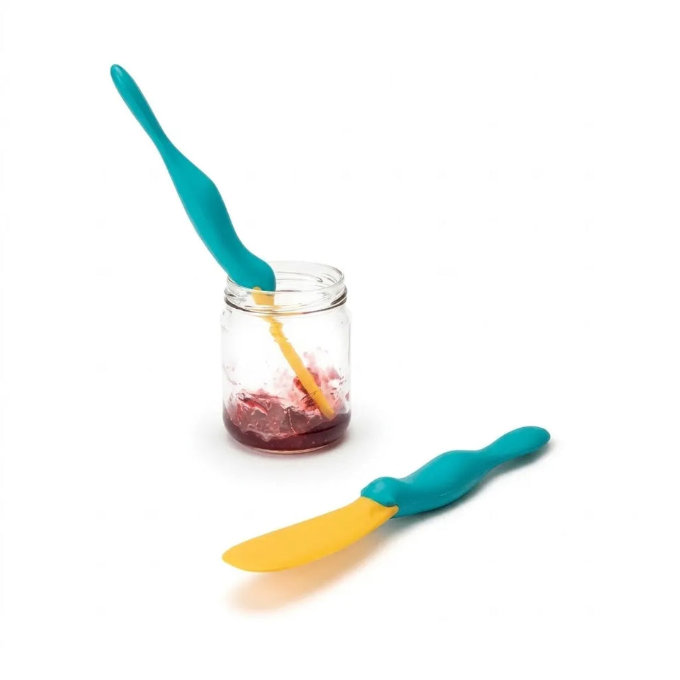 Two silicone spreader tools with teal handles and yellow blades, one resting inside a clear glass jar and one placed outside, on a white background.