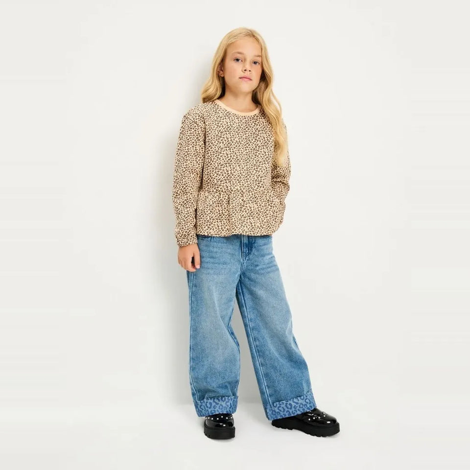 A young girl with blonde hair stands against a plain white studio background wearing a beige and brown patterned knit long-sleeve top, wide-leg blue jeans, and black shoes.