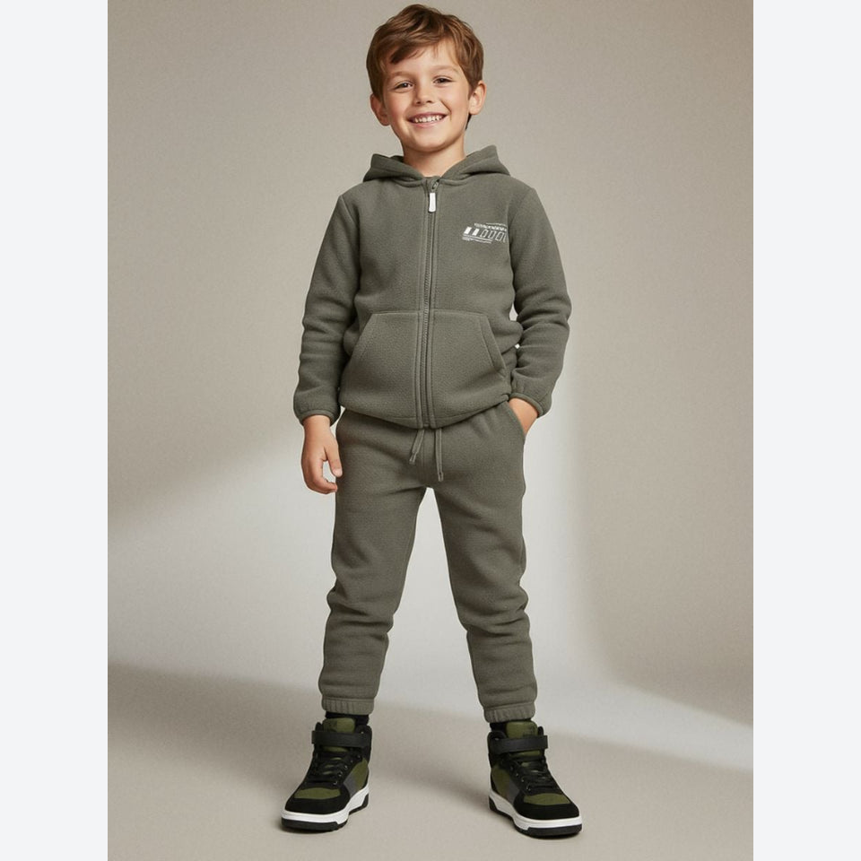 A young boy stands against a neutral studio backdrop wearing a matching olive green zip-up hoodie and jogger pants set, paired with black and green sneakers.