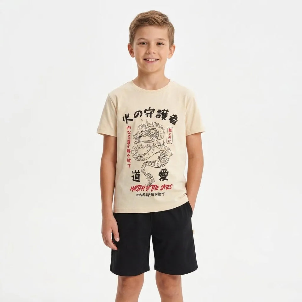 A young boy modeling a beige graphic t-shirt with a black dragon design and black shorts, standing against a plain white background.
