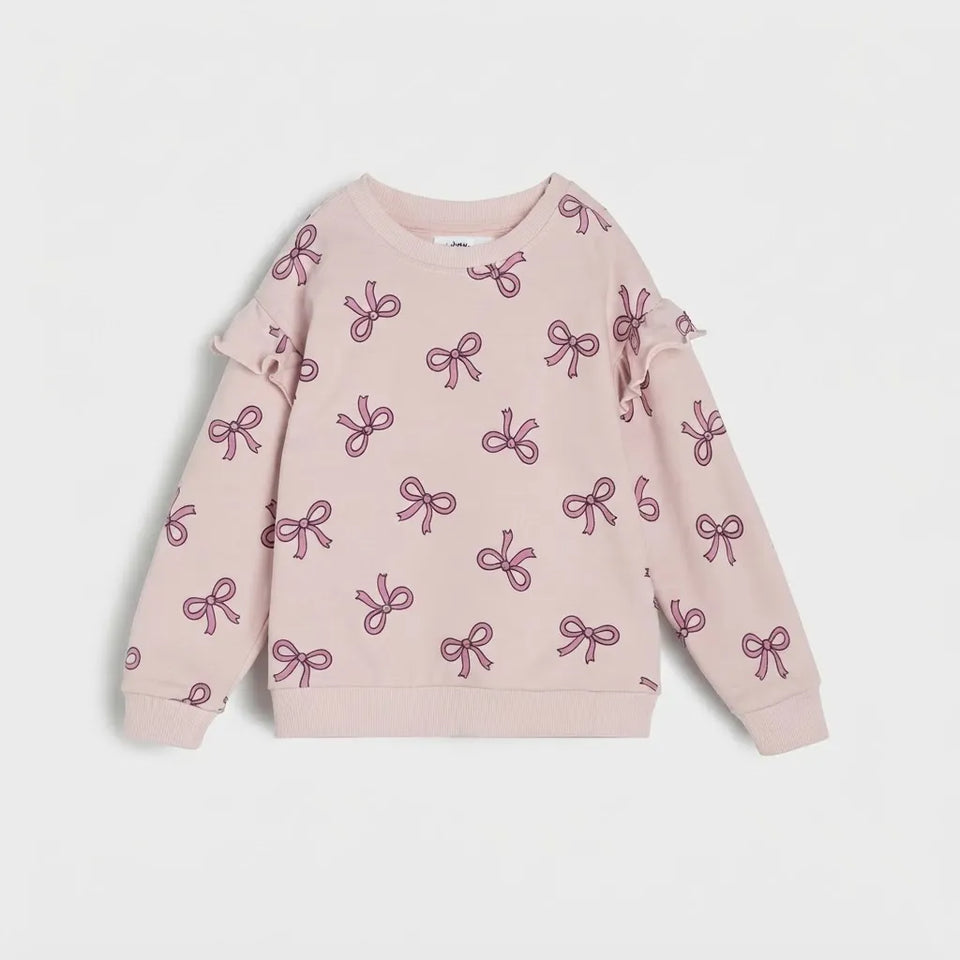 A light pink long-sleeve sweatshirt for toddlers, featuring an all-over pattern of darker pink bows and ruffle details on the shoulders, displayed against a light gray background.